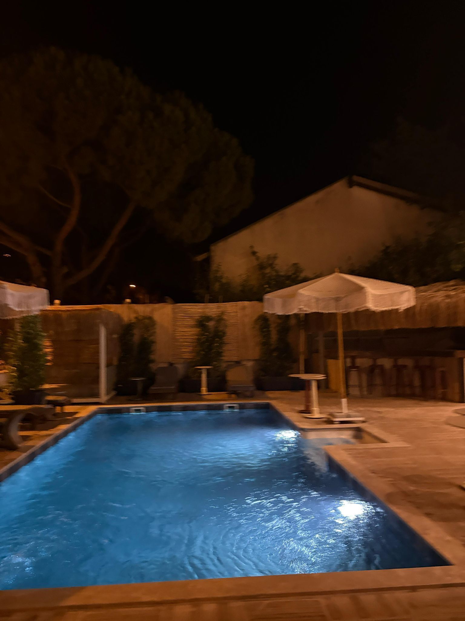 A swimming pool with a wooden deck, surrounded by umbrellas and potted plants.