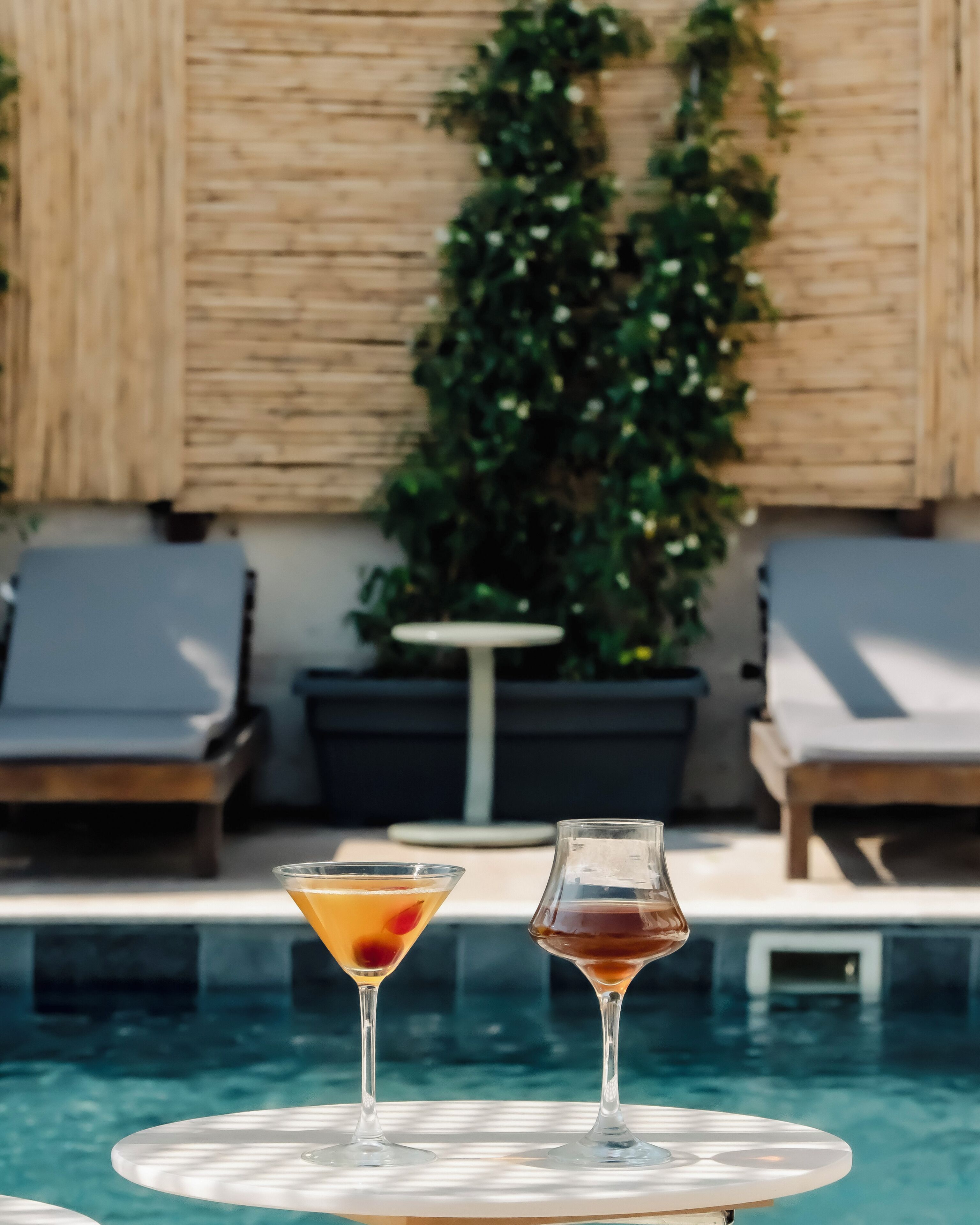 Une scène au bord de la piscine, avec deux boissons sur une table, entourée de chaises longues et d’un mur en bois agrémenté de verdure.