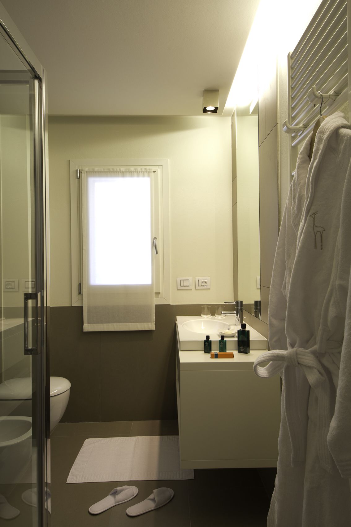 A bathroom with a glass shower enclosure, a toilet, a sink with toiletries, a towel rack, and a window with curtains.