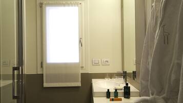 A bathroom with a glass shower enclosure, a toilet, a sink with toiletries, a towel rack, and a window with curtains.