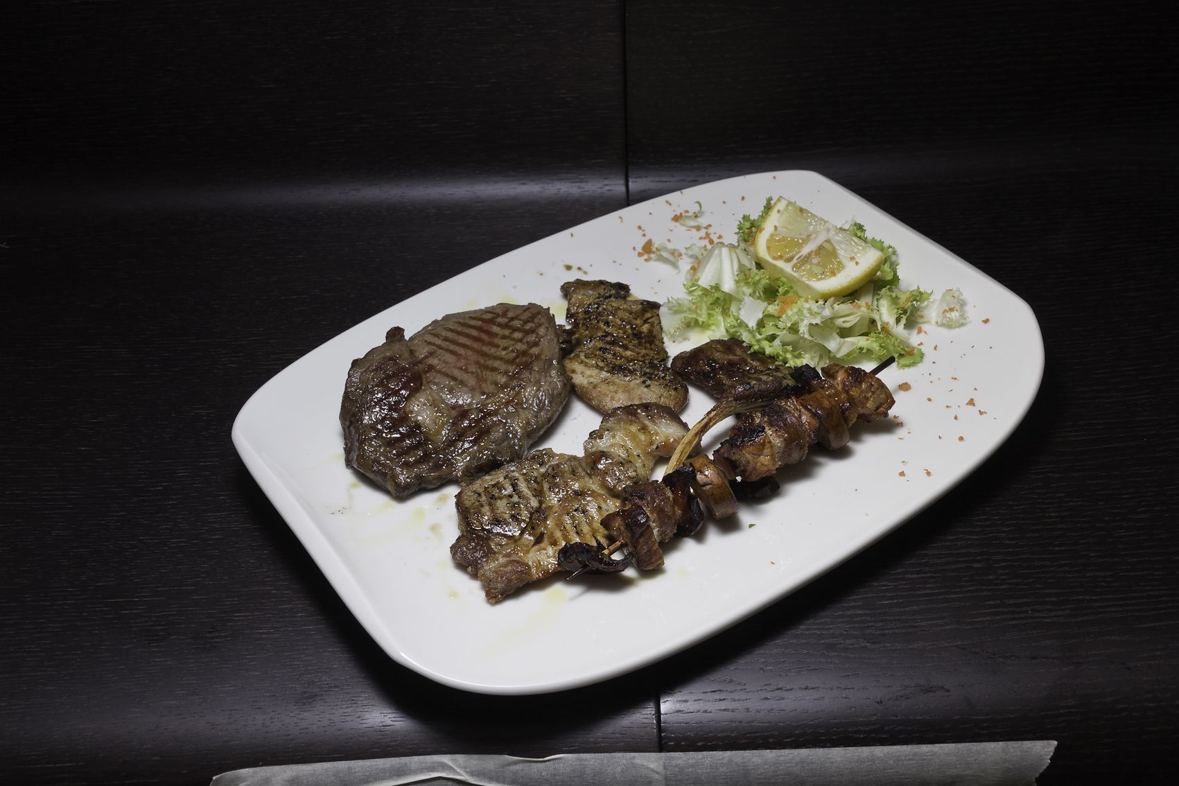 A plate of grilled meat with a side of lettuce and a slice of lemon on a white plate.