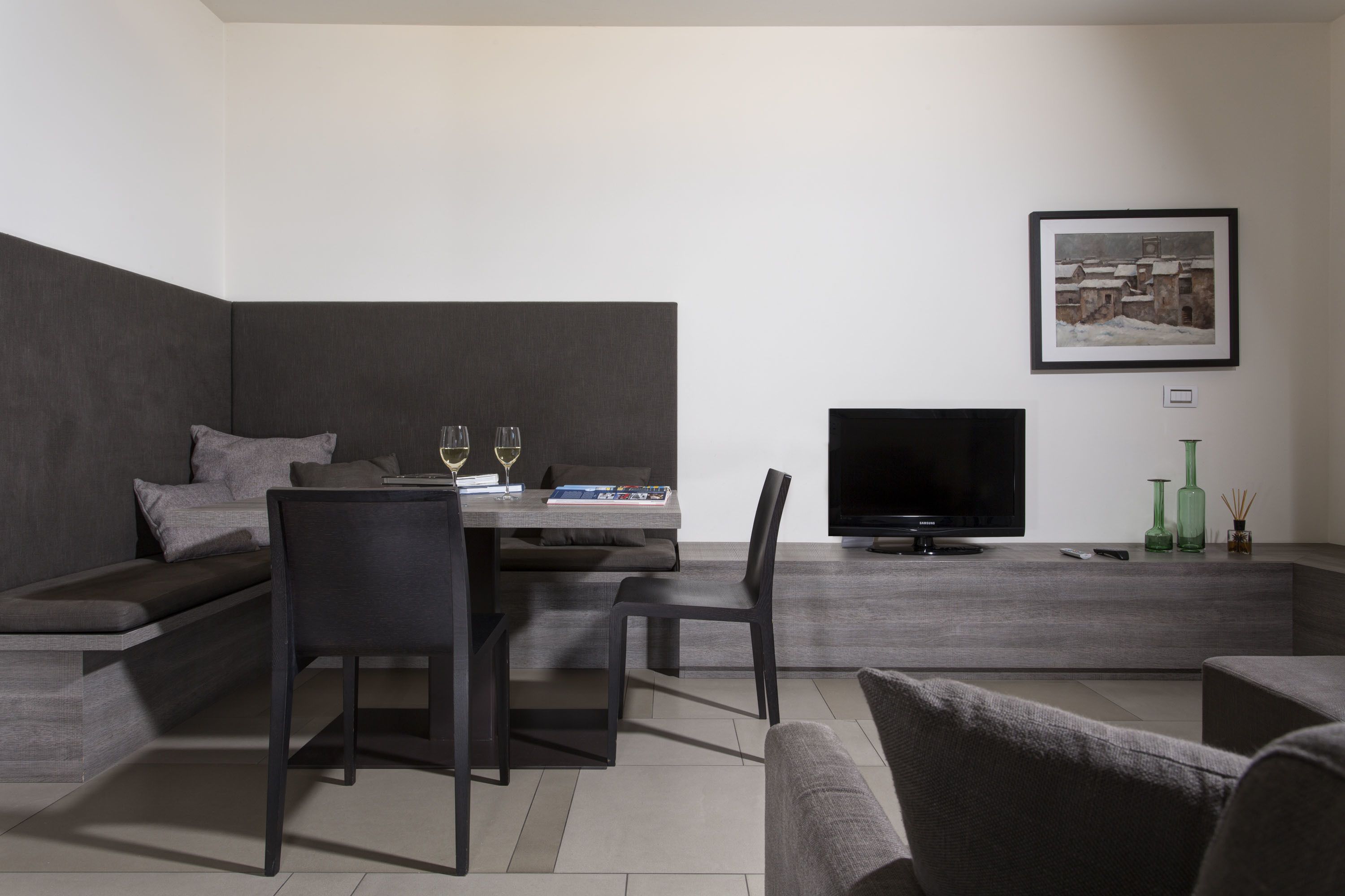 A modern living room with a sofa, dining table, and chairs. A flat-screen TV is mounted on the wall.