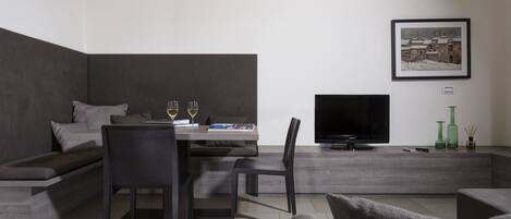A modern living room with a sofa, dining table, and chairs. A flat-screen TV is mounted on the wall.