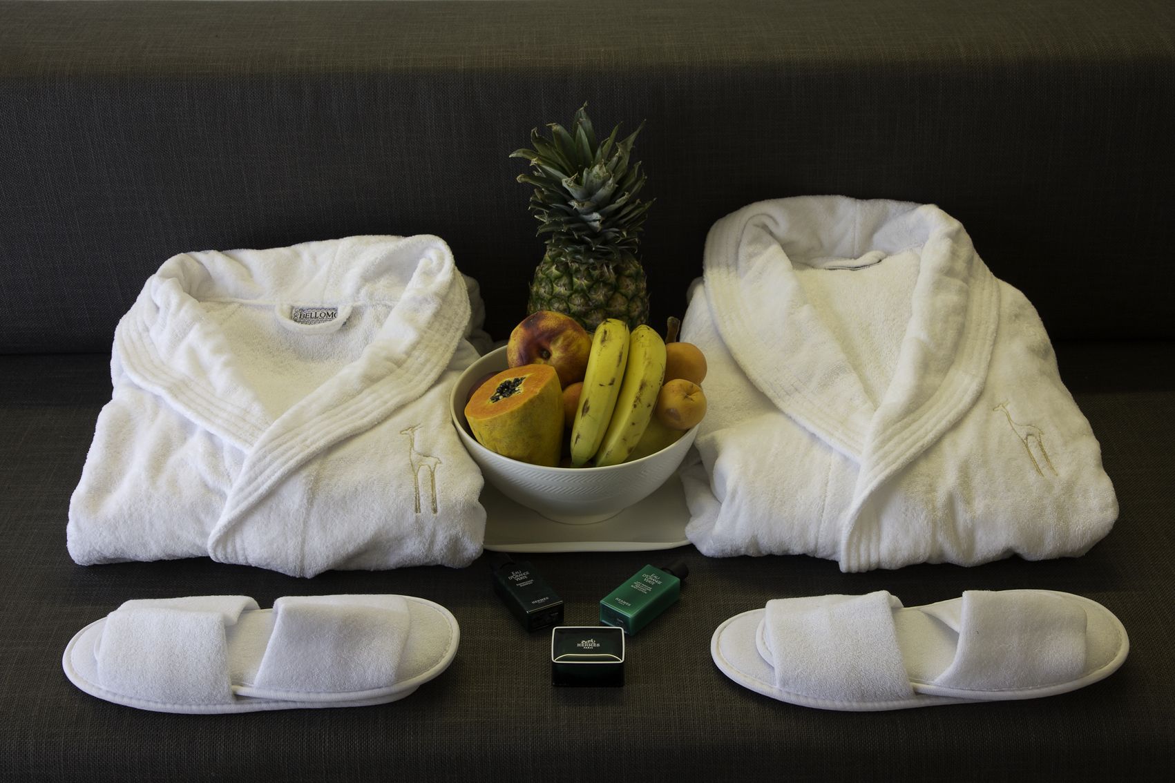 A room with a dark sofa, two white towels, a bowl of fruit, and slippers.