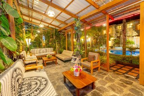 Un espacio al aire libre cubierto con mobiliario de madera, suelo de piedra y abundante vegetación.