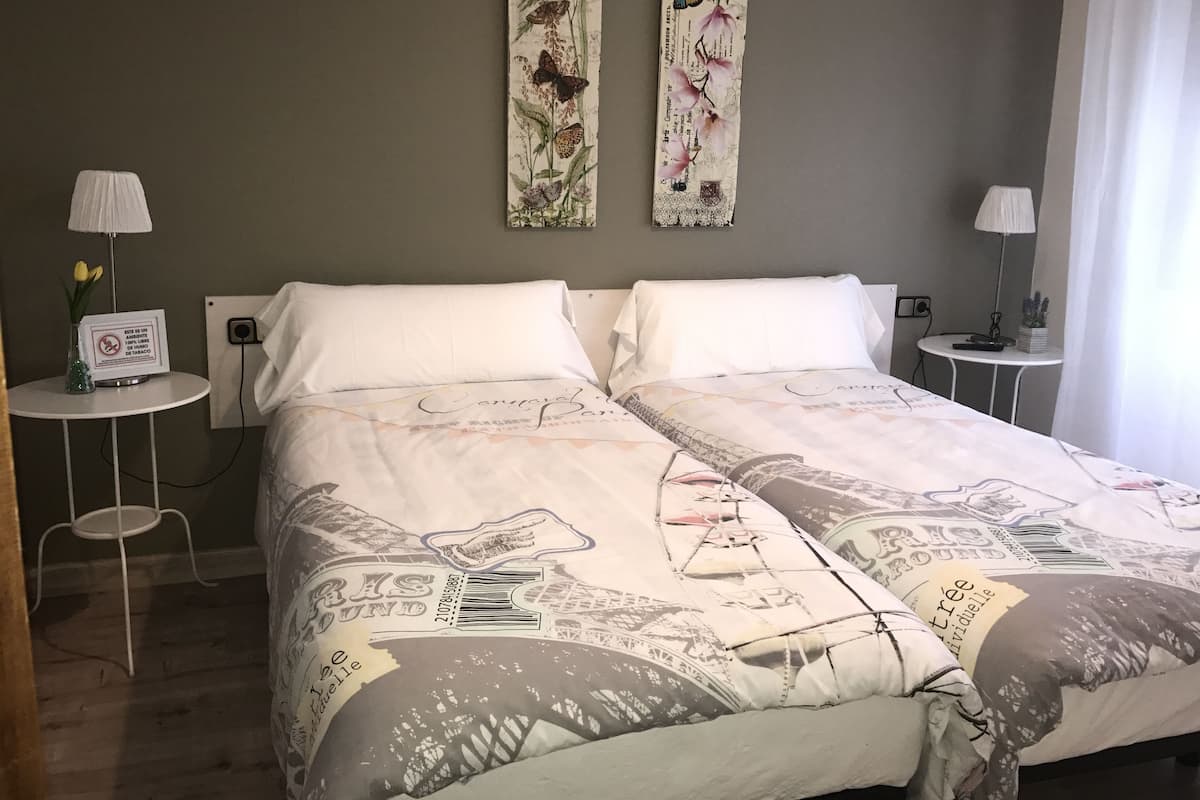 En dubbelsäng med ett mönstrat överkast, två sängbord med lampor, ett vägguttag och ett litet bord med en vas med blommor.