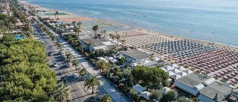 An aerial view of a beachside resort with rows of beach umbrellas, a parking lot, and a road lined with palm trees.