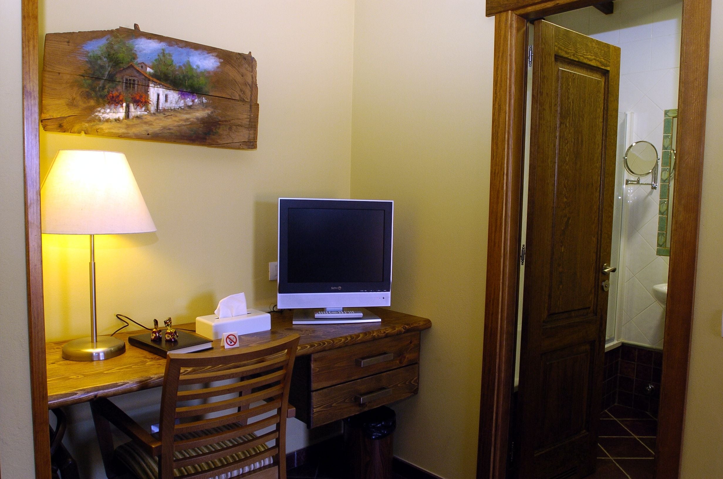 Un escritorio pequeño con una computadora, una lámpara y una silla. Se ve una puerta de madera y un baño al fondo.