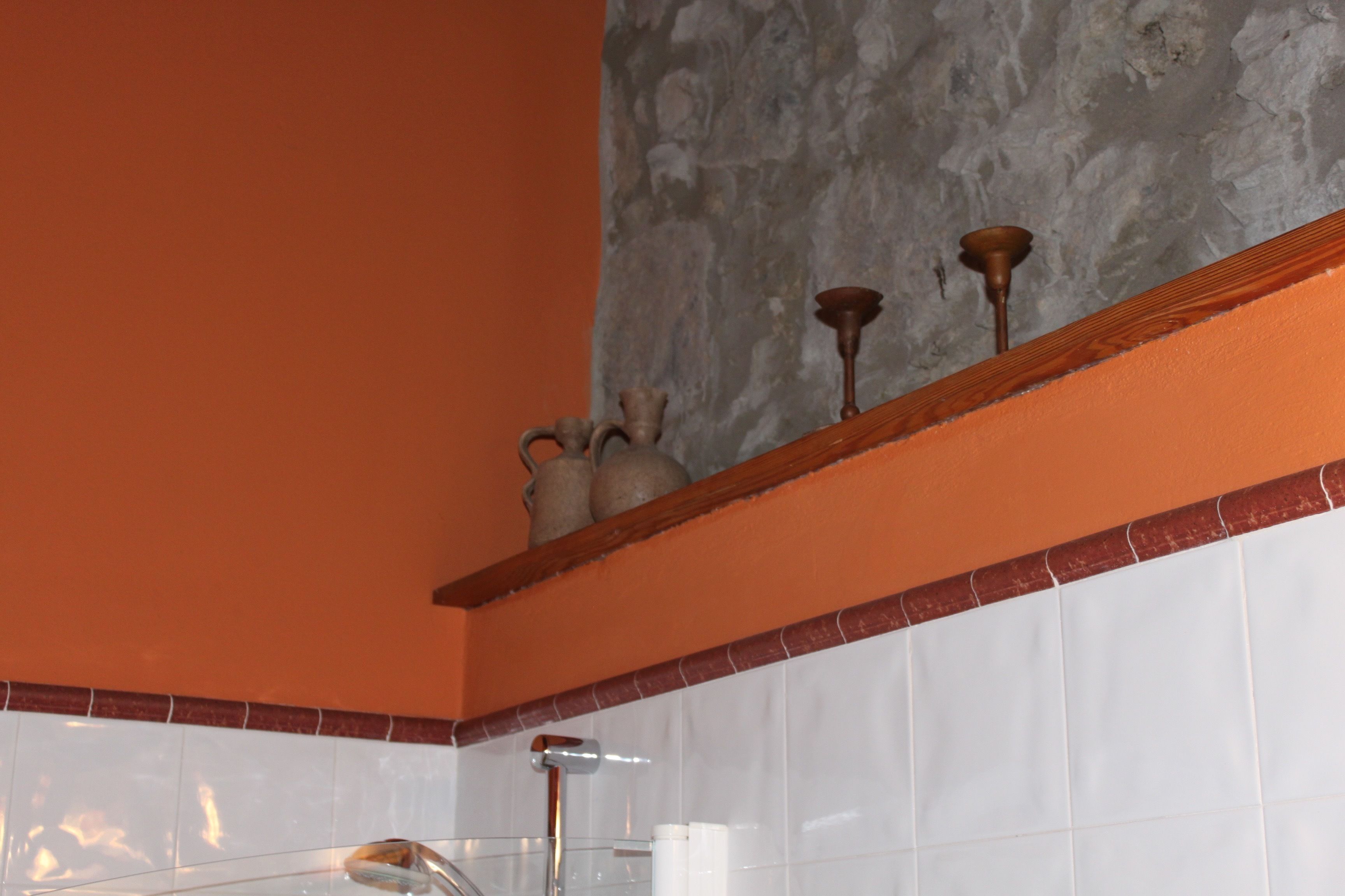 A bathroom with a white tiled wall, an orange accent wall, and a wooden shelf with decorative items.