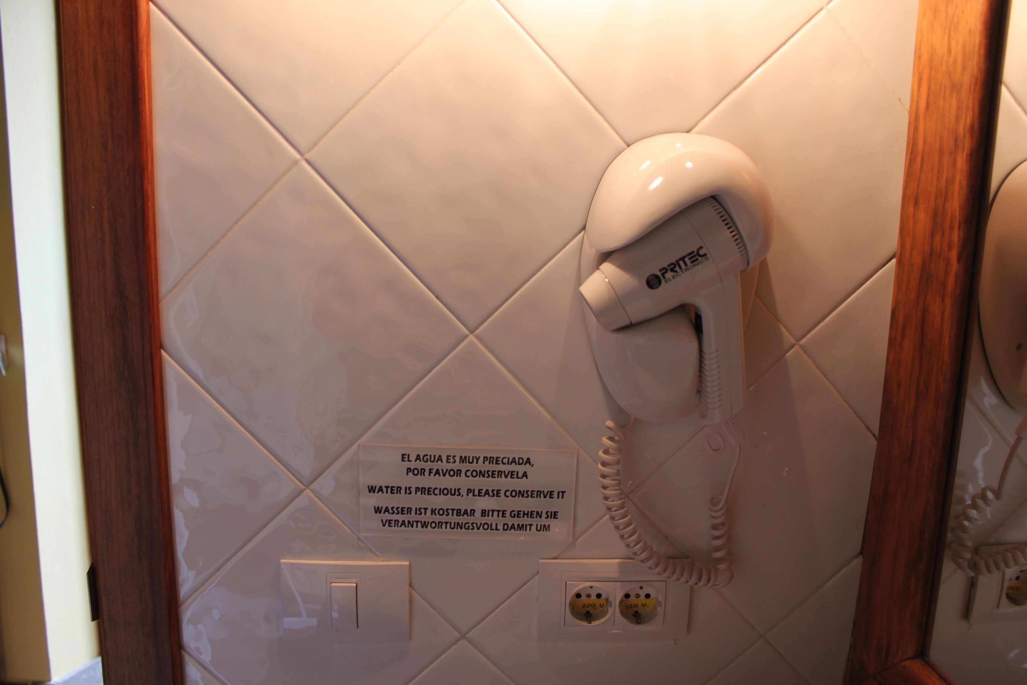 Une salle de bain avec un téléphone blanc, un panneau invitant à économiser l’eau et des prises électriques.