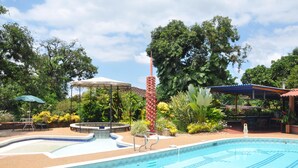 En swimmingpool med fliseterrasse, omgivet af frodig vegetation og skyggefulde områder med parasoller.