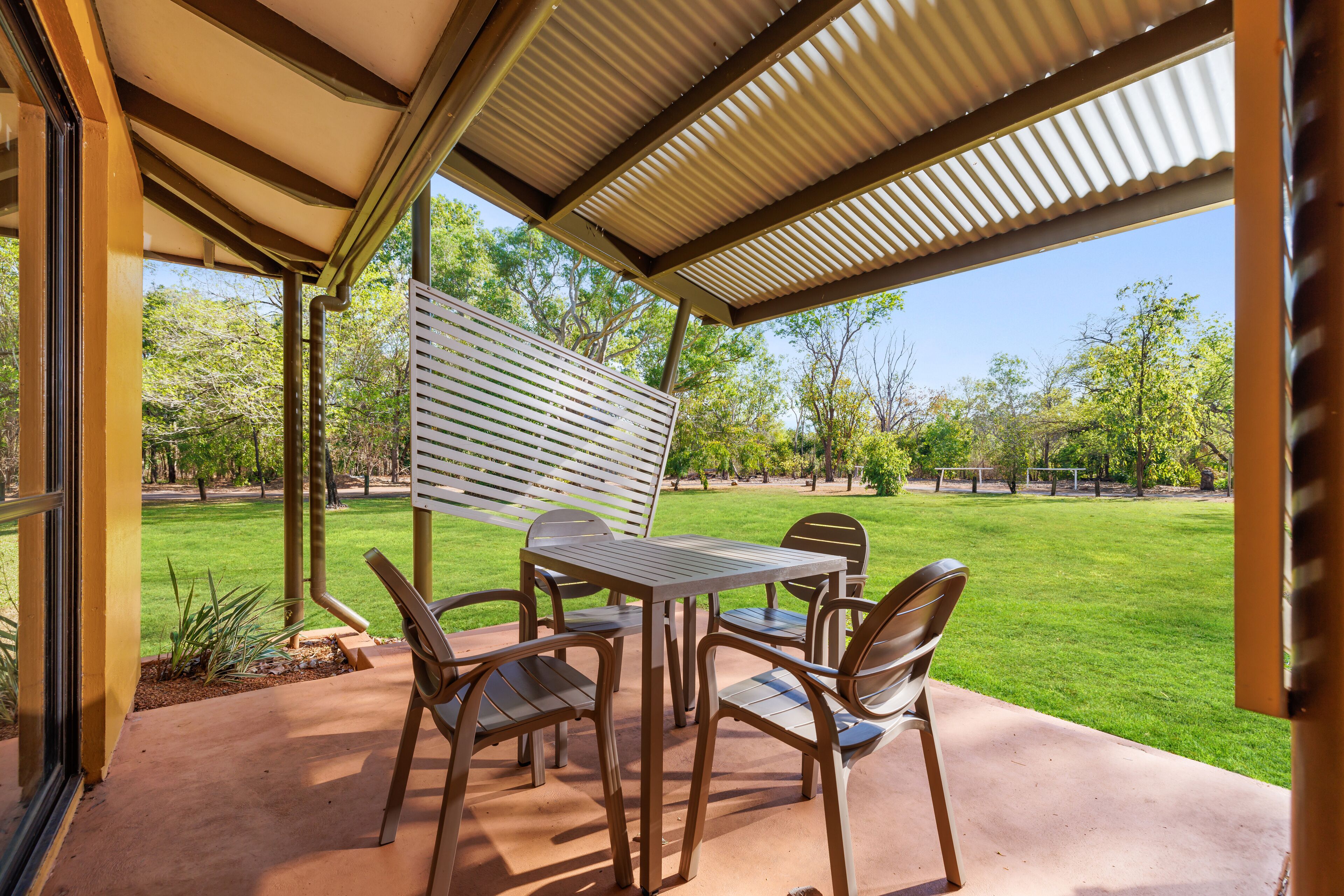 Eine Terrasse im Freien mit Tisch und Stühlen, einer Pergola darüber und Blick auf einen gepflegten Rasen und Bäume.