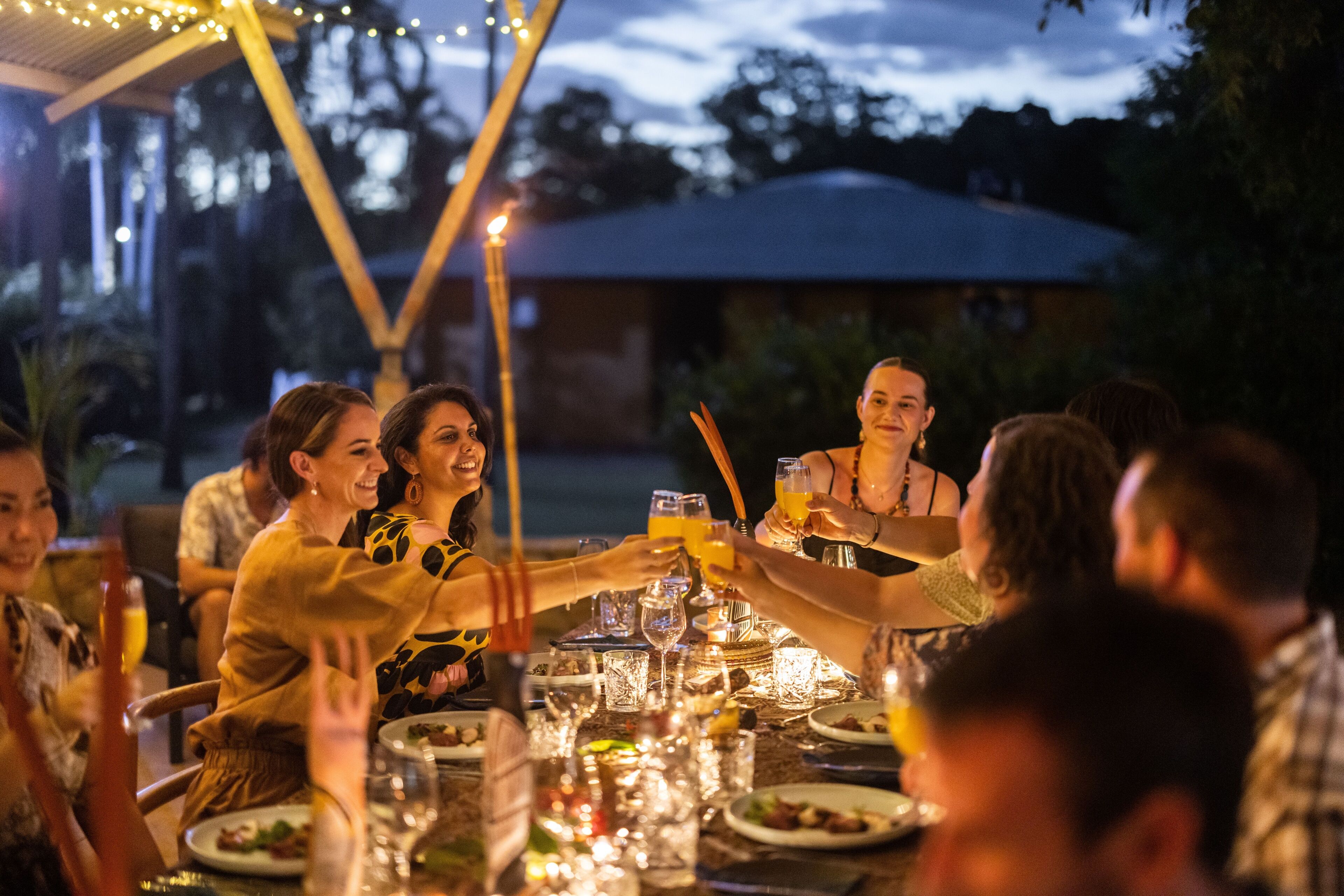 A group of people toasting at an outdoor dining table with candles and drinks, surrounded by a covered area with string lights.