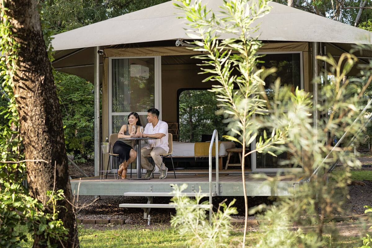 Un couple est assis à une table, à l’abri sous une pergola, avec vue sur une zone boisée.