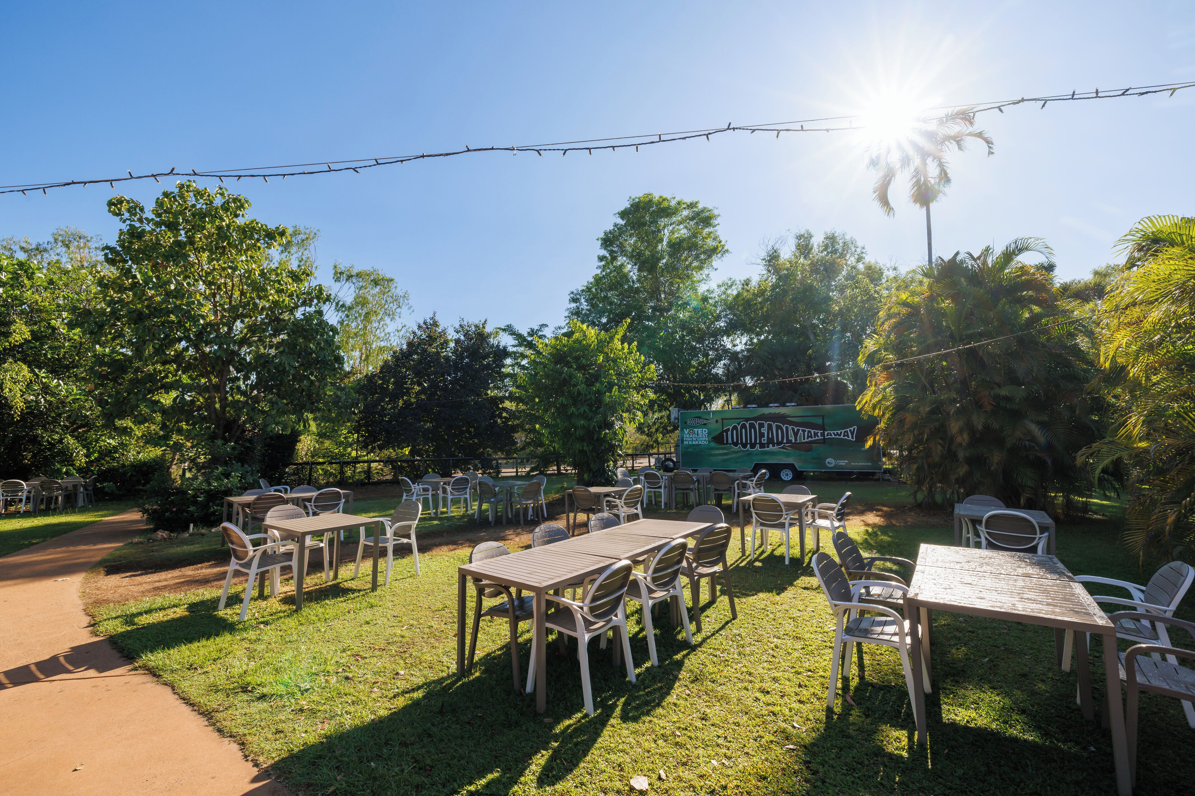 An outdoor seating area with tables and chairs, surrounded by trees and a green food truck in the background.