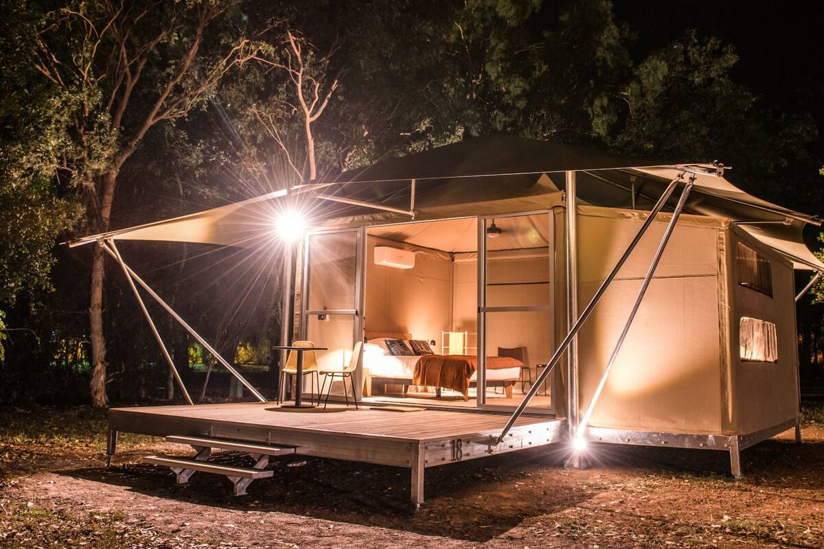 Une tente spacieuse et bien éclairée, dotée d’une terrasse en bois, d’un lit et d’un coin salon, située dans une zone boisée.