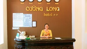 Un mostrador de recepción de hotel con relojes que muestran diferentes ciudades, una recepcionista y carteles informativos en la pared.