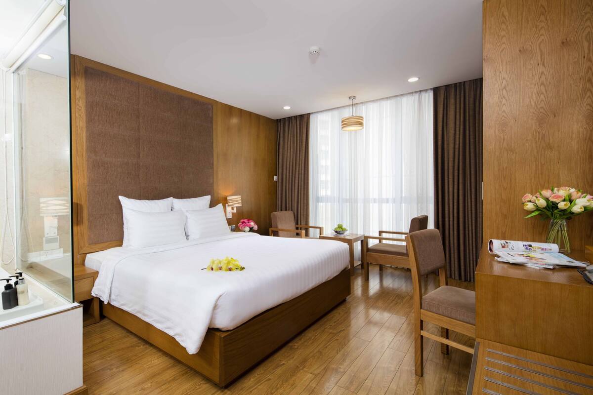A hotel room with a large bed, a desk with a chair, a small table with a vase of flowers, and a window with curtains.