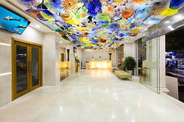 Un vestíbulo de hotel con un mostrador de recepción, un amplio muro de vidrio y un techo decorado con arte de vidrio de colores.