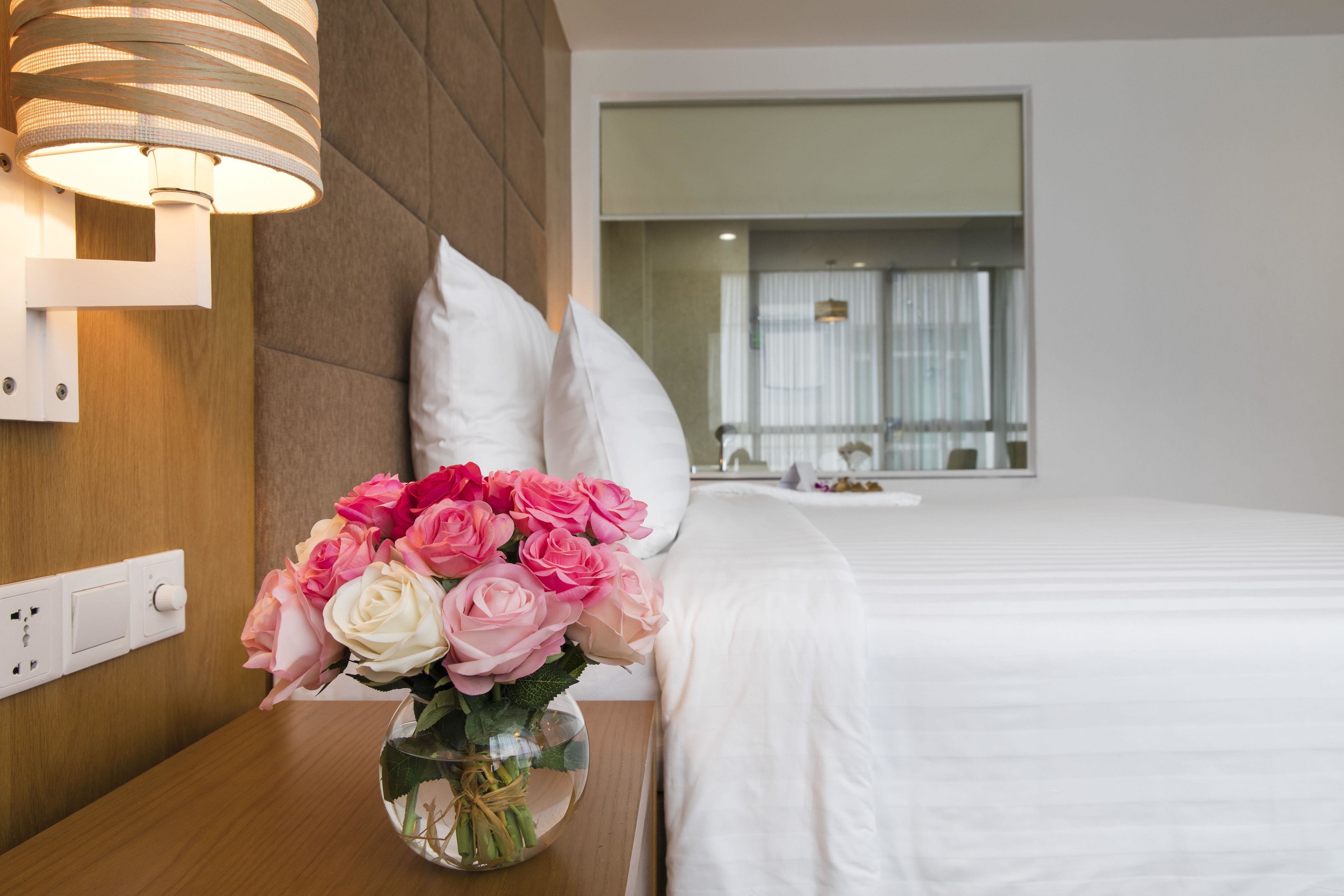 Ein Hotelzimmer mit einem Bett, einem Nachttisch mit einer Vase voller pinkfarbener Rosen, einer Wandleuchte und einem Fenster mit Vorhängen.