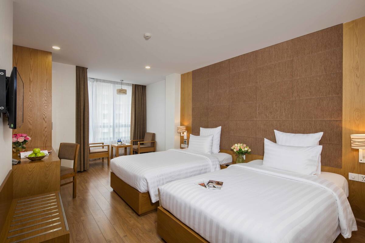 A hotel room with two beds, a desk with a chair, a small table with a vase of flowers, and a television on the wall.