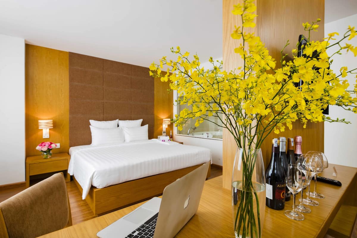A hotel room with a bed, bedside tables, a desk with a laptop, a vase with flowers, and wine bottles.