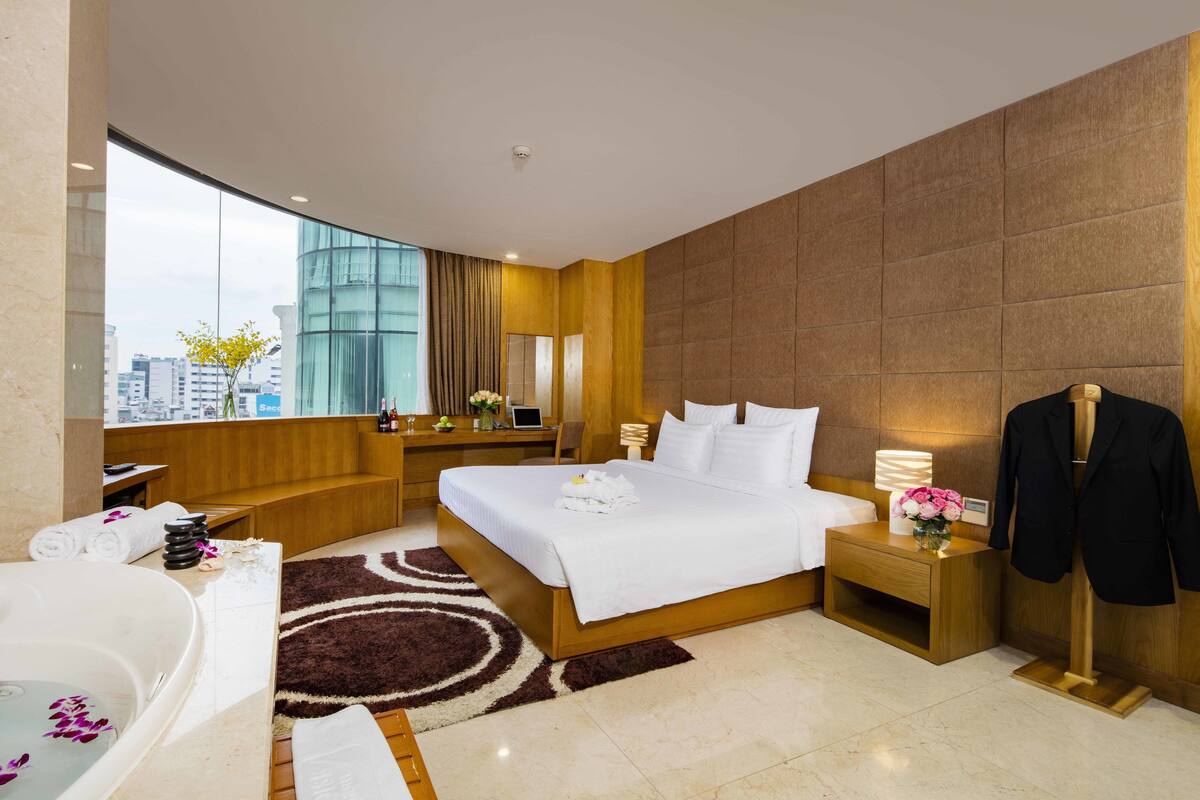 A modern hotel room with a large bed, a sitting area, a desk, and a view of the city.