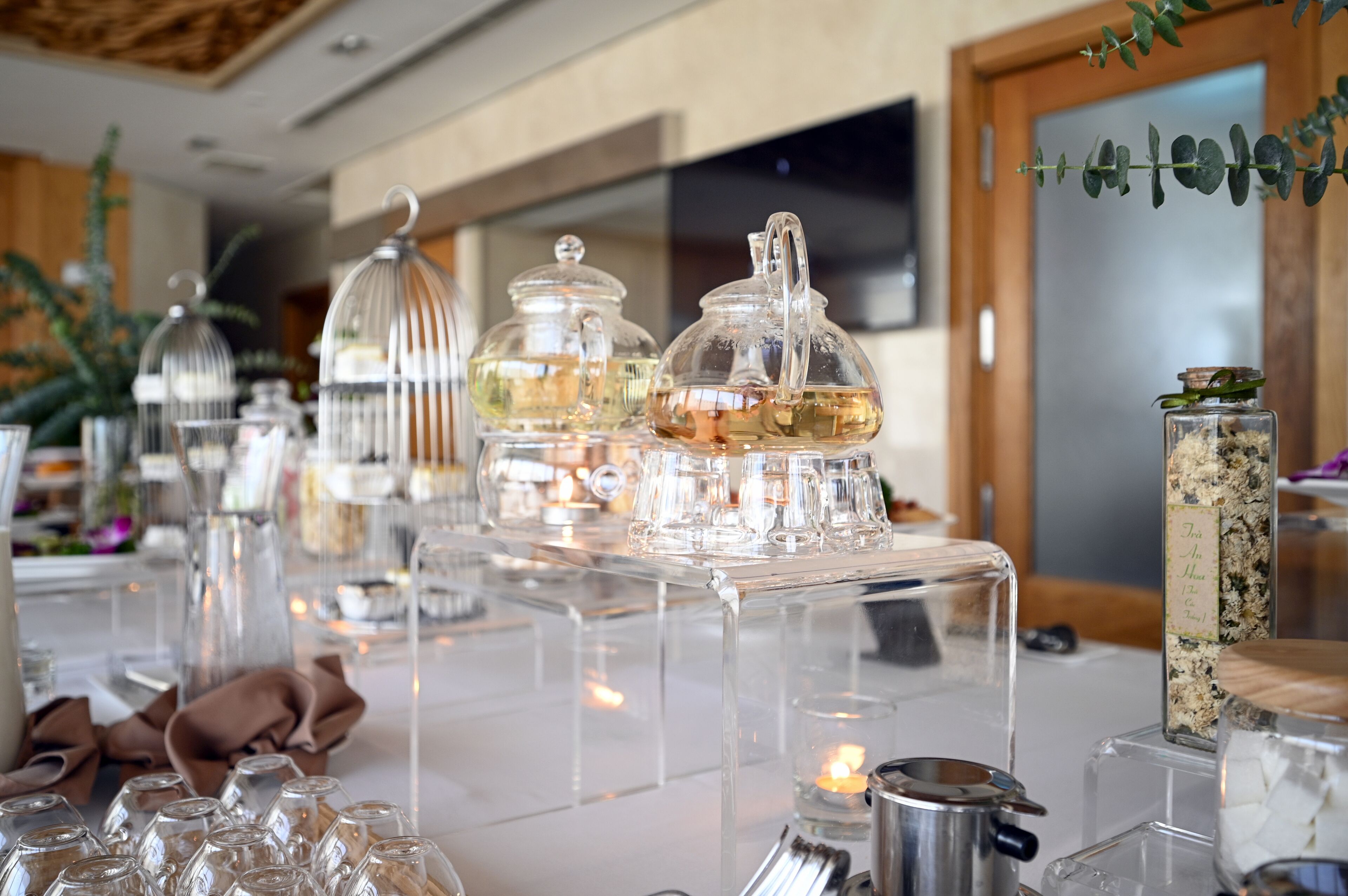 Une table surmontée de récipients en verre transparent et d’un chandelier, placée dans une pièce bien éclairée, avec une porte en bois et une plante en arrière-plan.