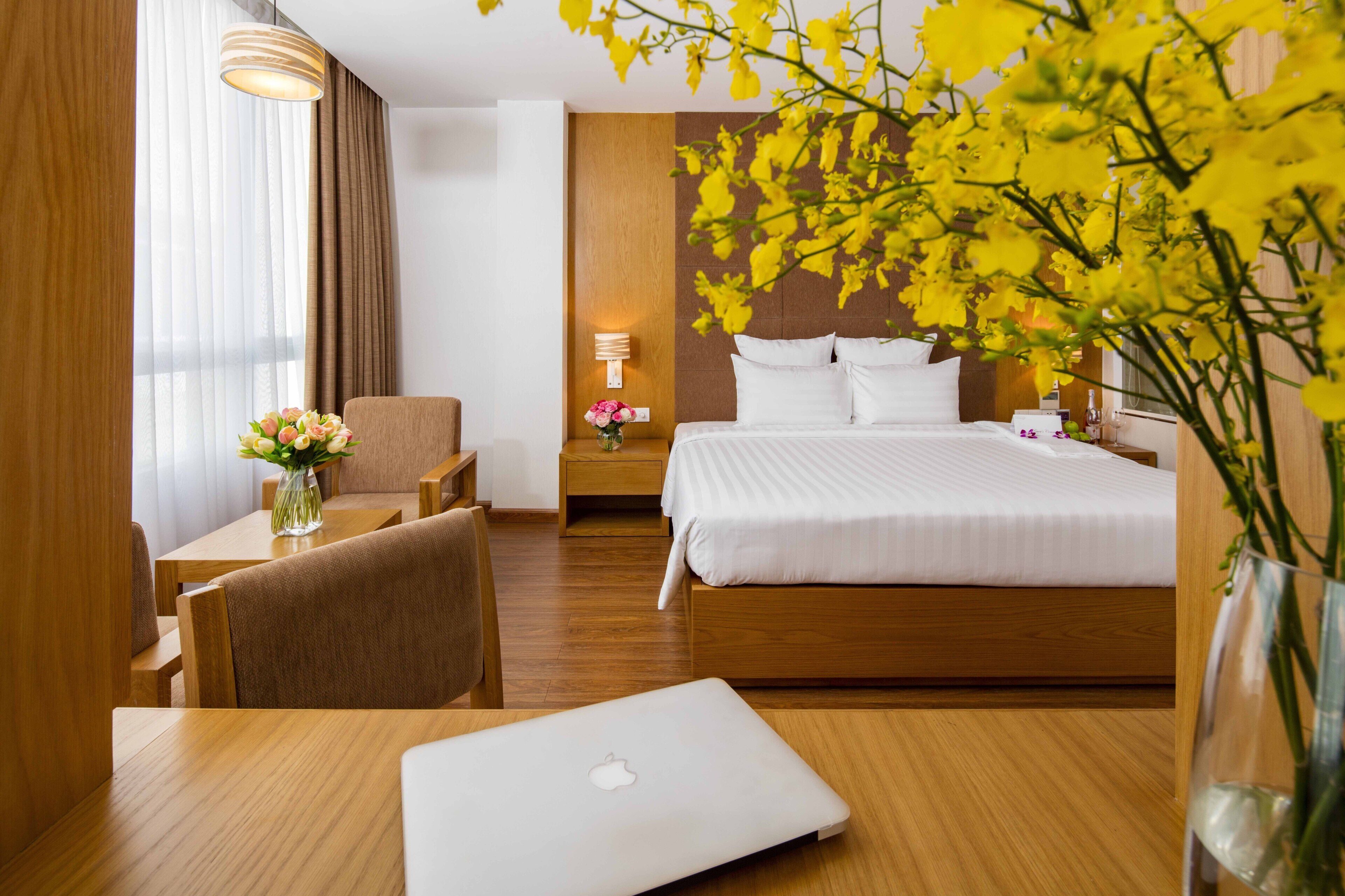 A hotel room with a bed, a desk with a laptop, a chair, a nightstand with a lamp, and a vase with flowers.