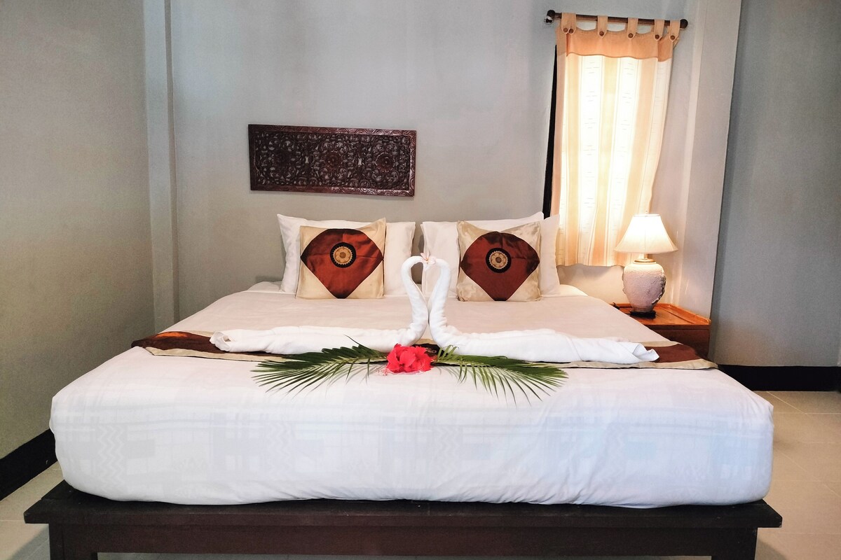 Een netjes opgemaakt bed met handdoeken gevouwen in de vorm van een zwaan, een decoratief kussen en een klein tafeltje met een lamp ernaast.