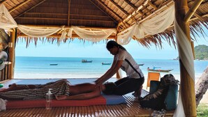 A beachside massage area with a thatched roof, a person receiving a massage, and a view of the ocean with boats. - Phuwadee Resort & Spa (Ko Pha-ngan)