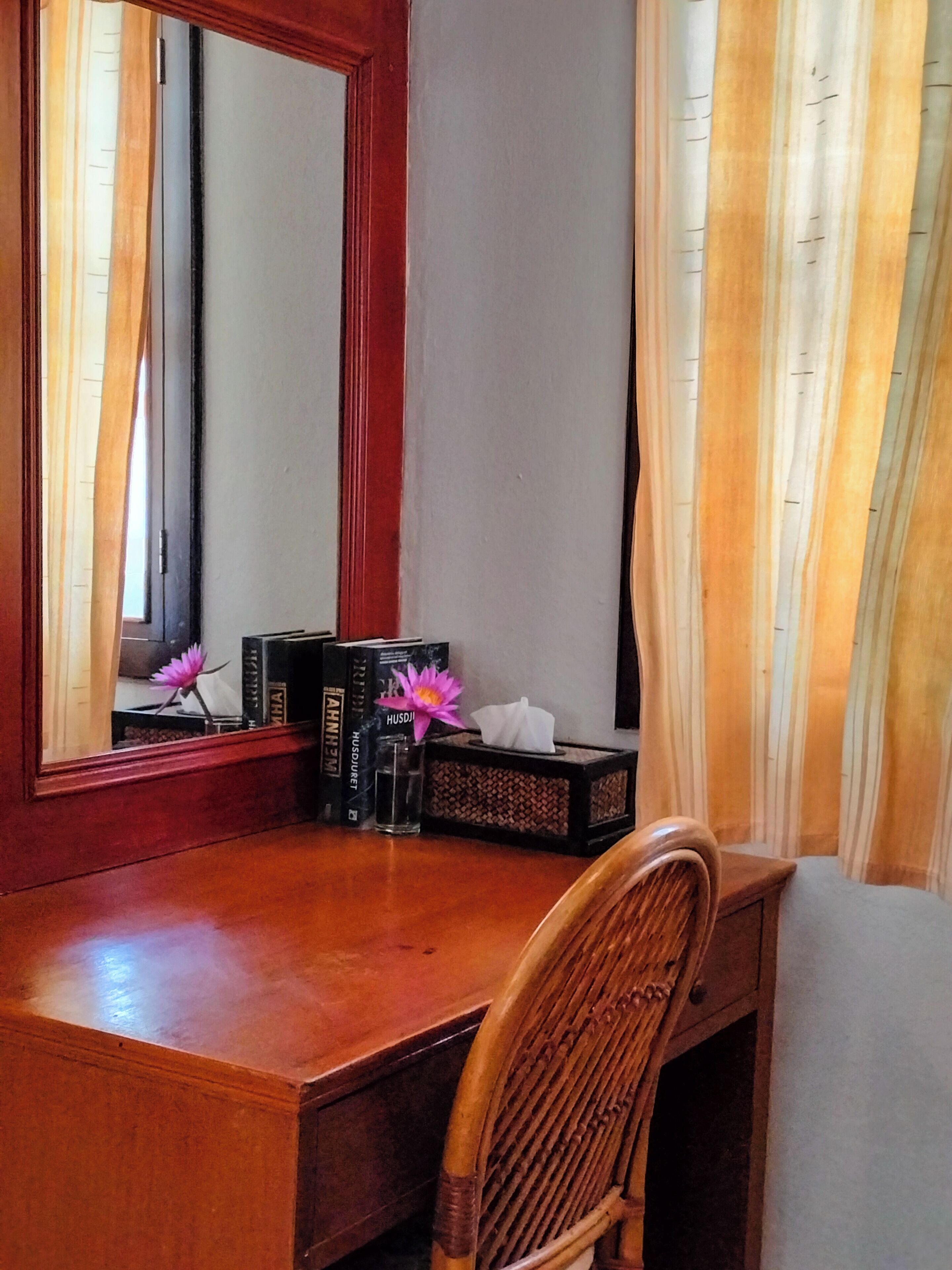 A wooden desk with a mirror, books, a tissue box, and a vase with flowers.
