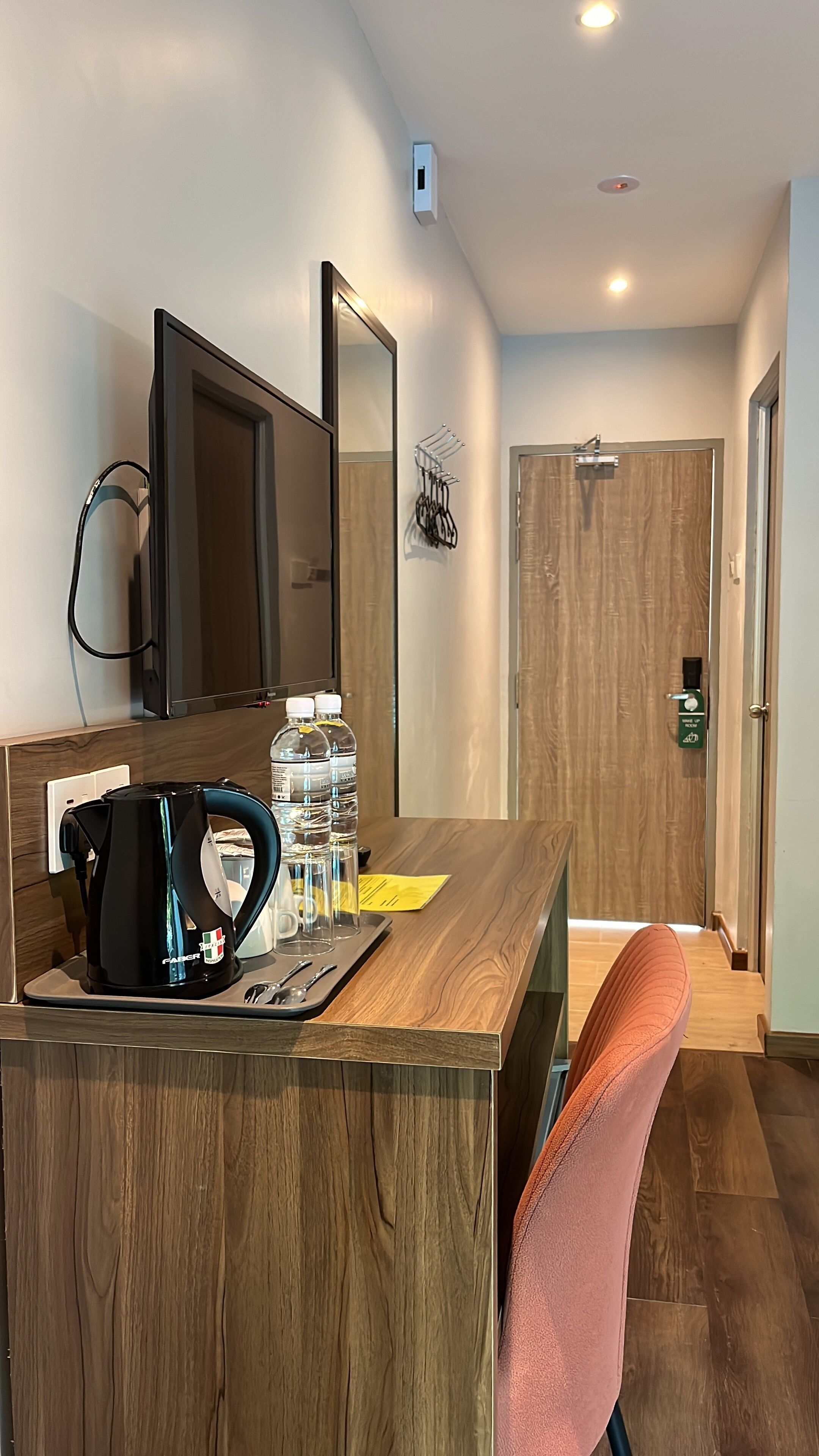 A modern hotel room with a wooden desk, a black kettle, water bottles, and a pink chair.