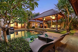 A poolside area with lounge chairs, a covered seating area, and a building with a tiled roof. - The Kampung Ubud Villa (Ubud)