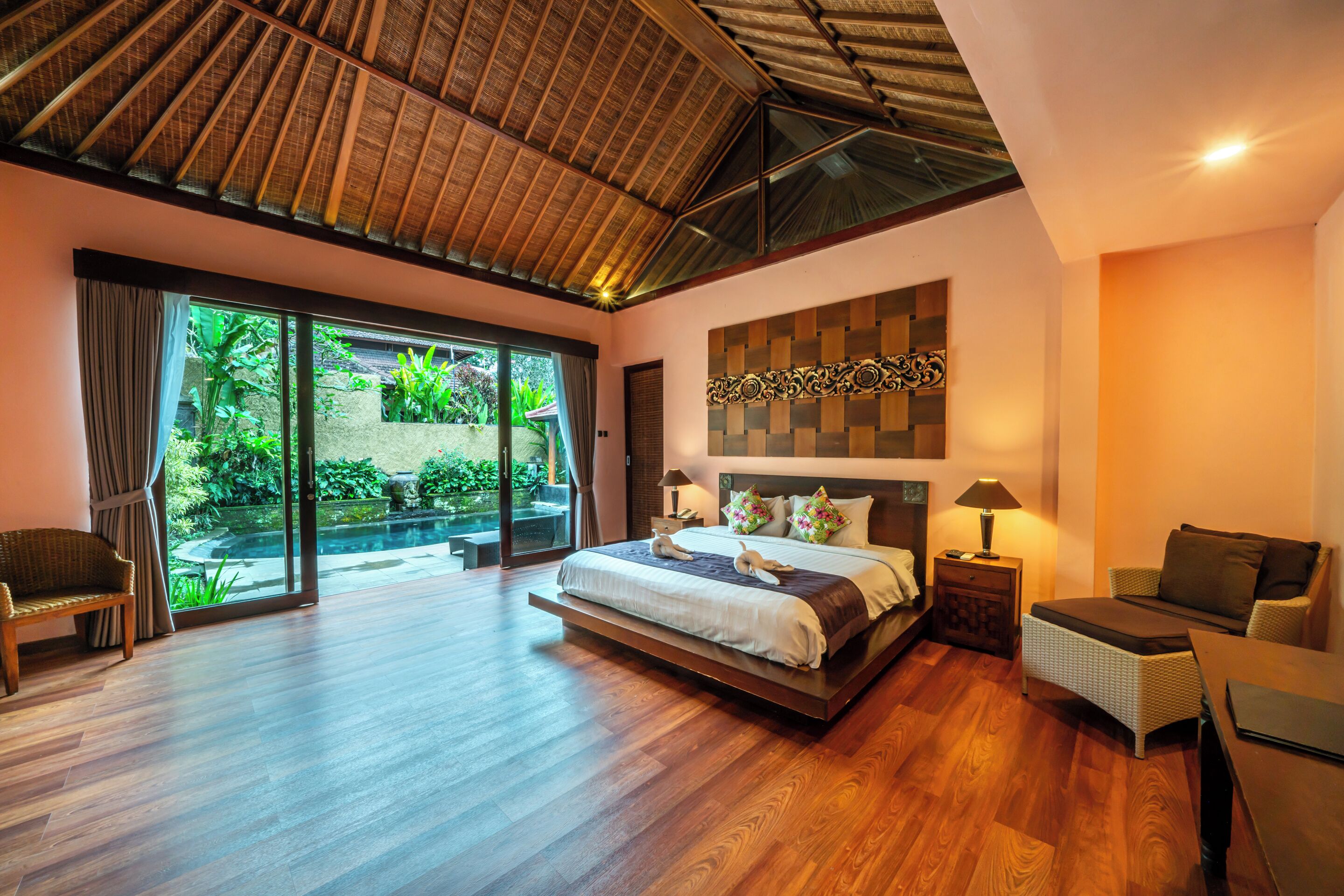 A spacious bedroom with a large bed, a wooden ceiling, and a view of a garden through a sliding glass door.