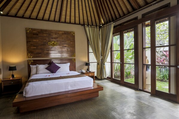 A large bed with a wooden headboard, two bedside tables, and a view of greenery through large windows. - The Kampung Ubud Villa (Ubud)