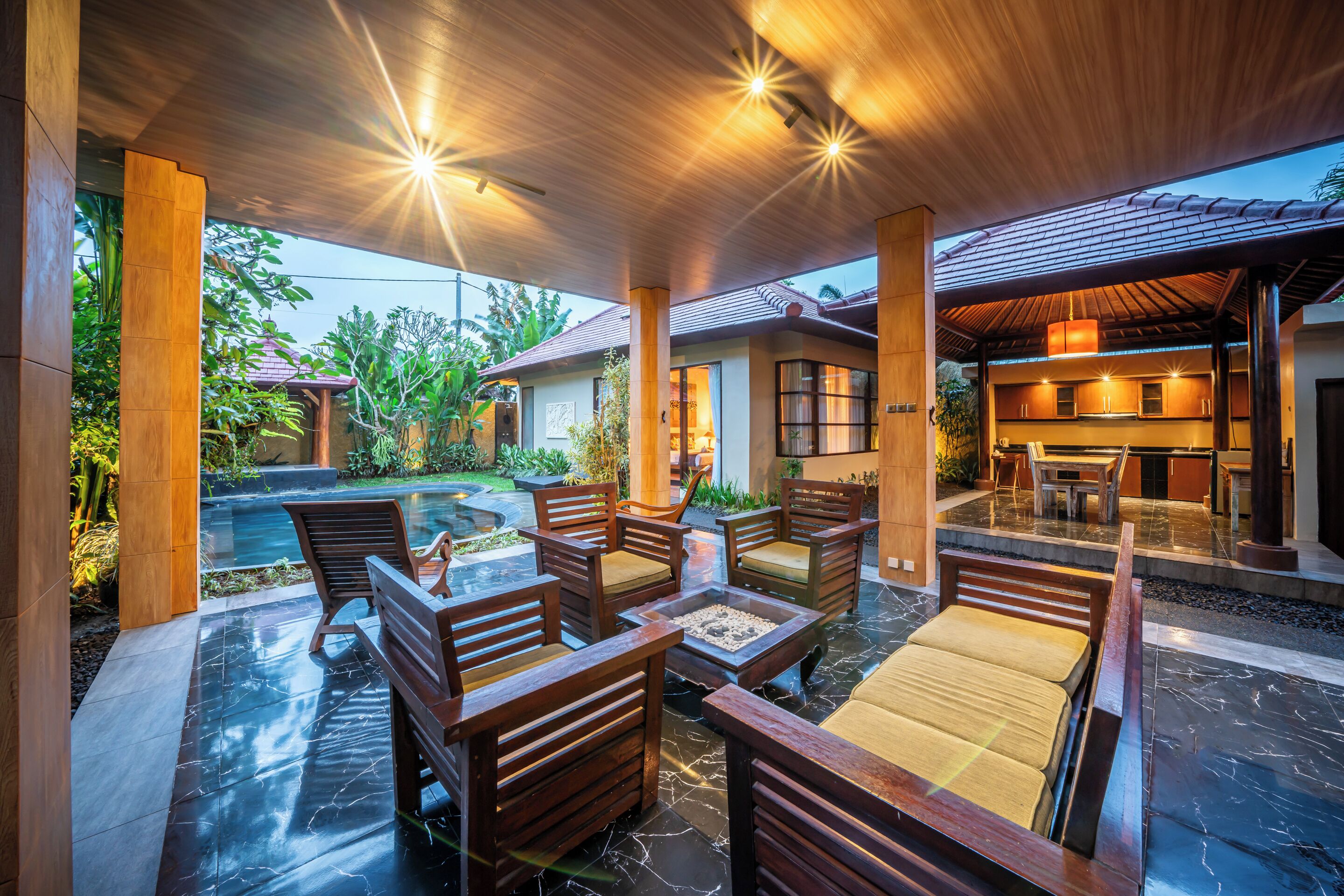 An outdoor seating area with wooden furniture and a fire pit, surrounded by a pool and lush greenery.