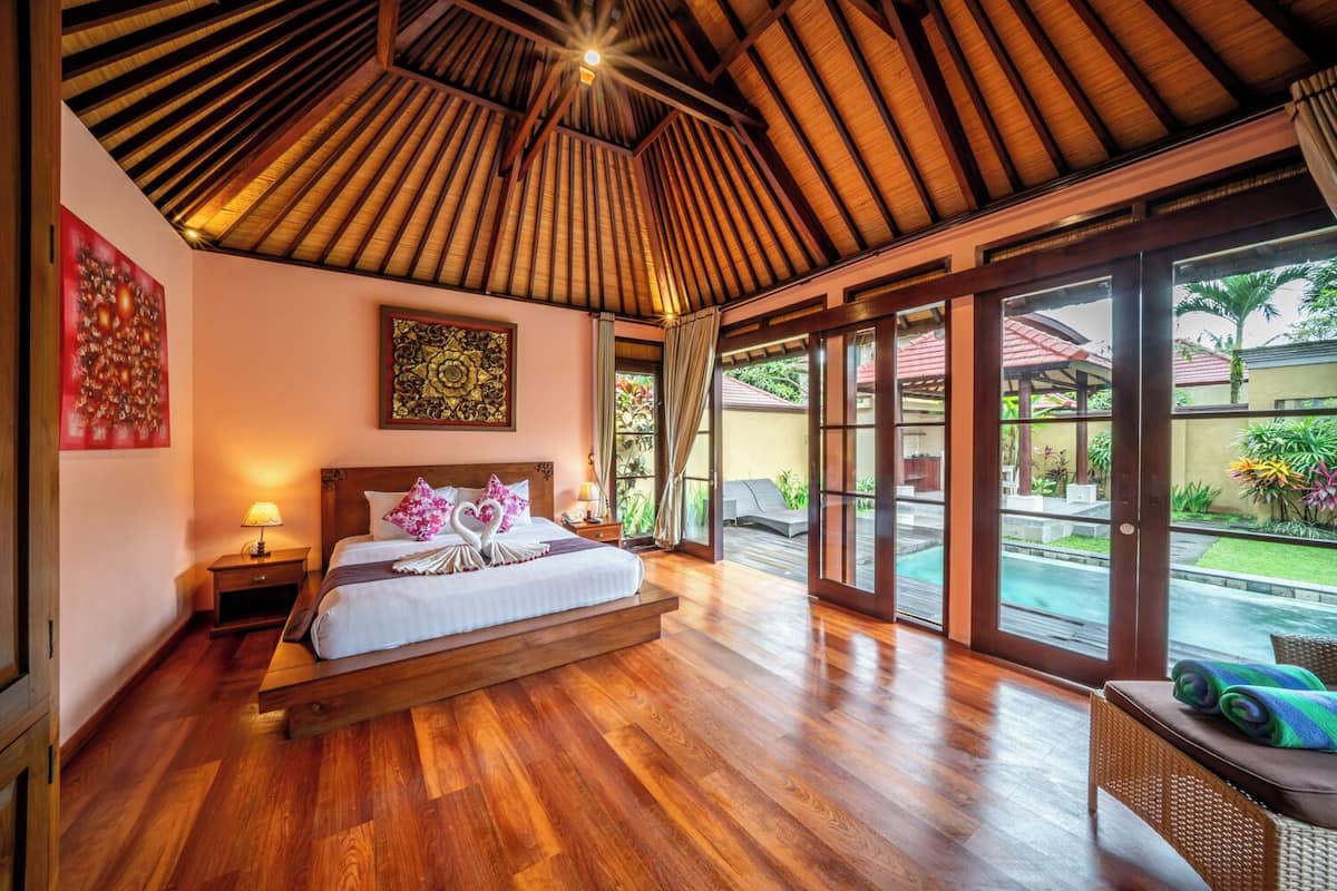 A spacious bedroom with a wooden ceiling, a large bed with floral pillows, a nightstand, and a view of a pool and garden through large sliding doors.