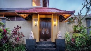 Entrada de um edifício com porta de madeira escura, duas luminárias na parede e uma placa sobre a porta.