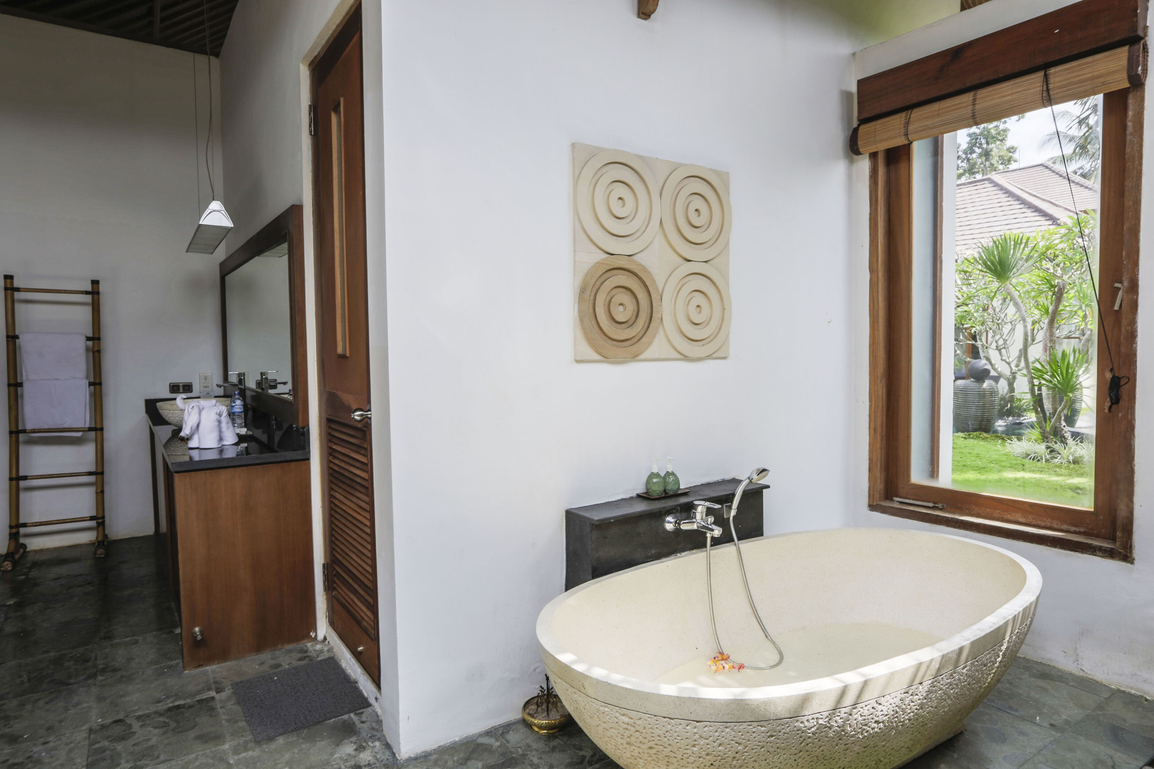 A bathroom with a large freestanding bathtub, a wooden vanity, a mirror, and a window with a view of greenery outside.