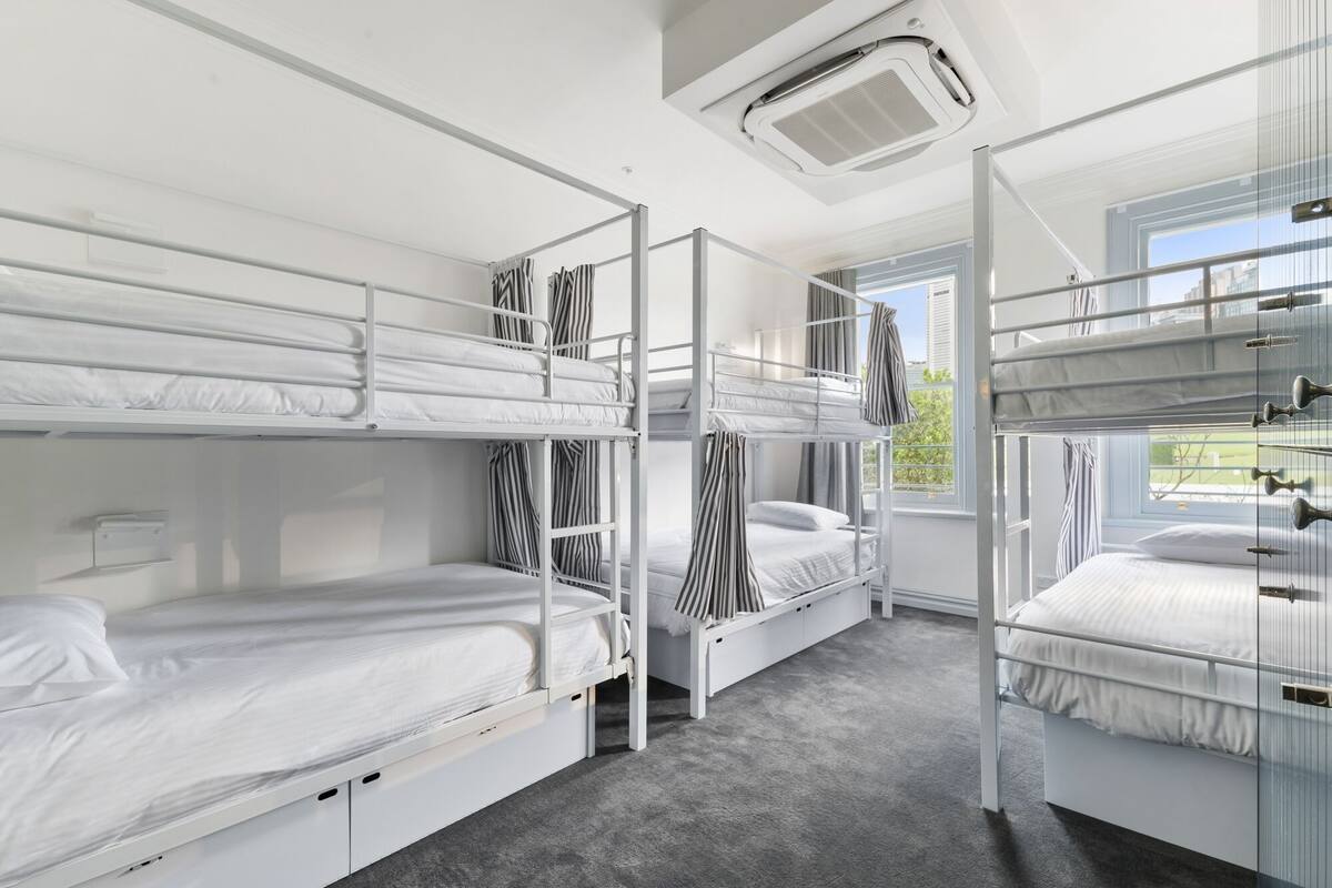 A dormitory room with bunk beds, a window with curtains, and an air conditioning unit on the ceiling.