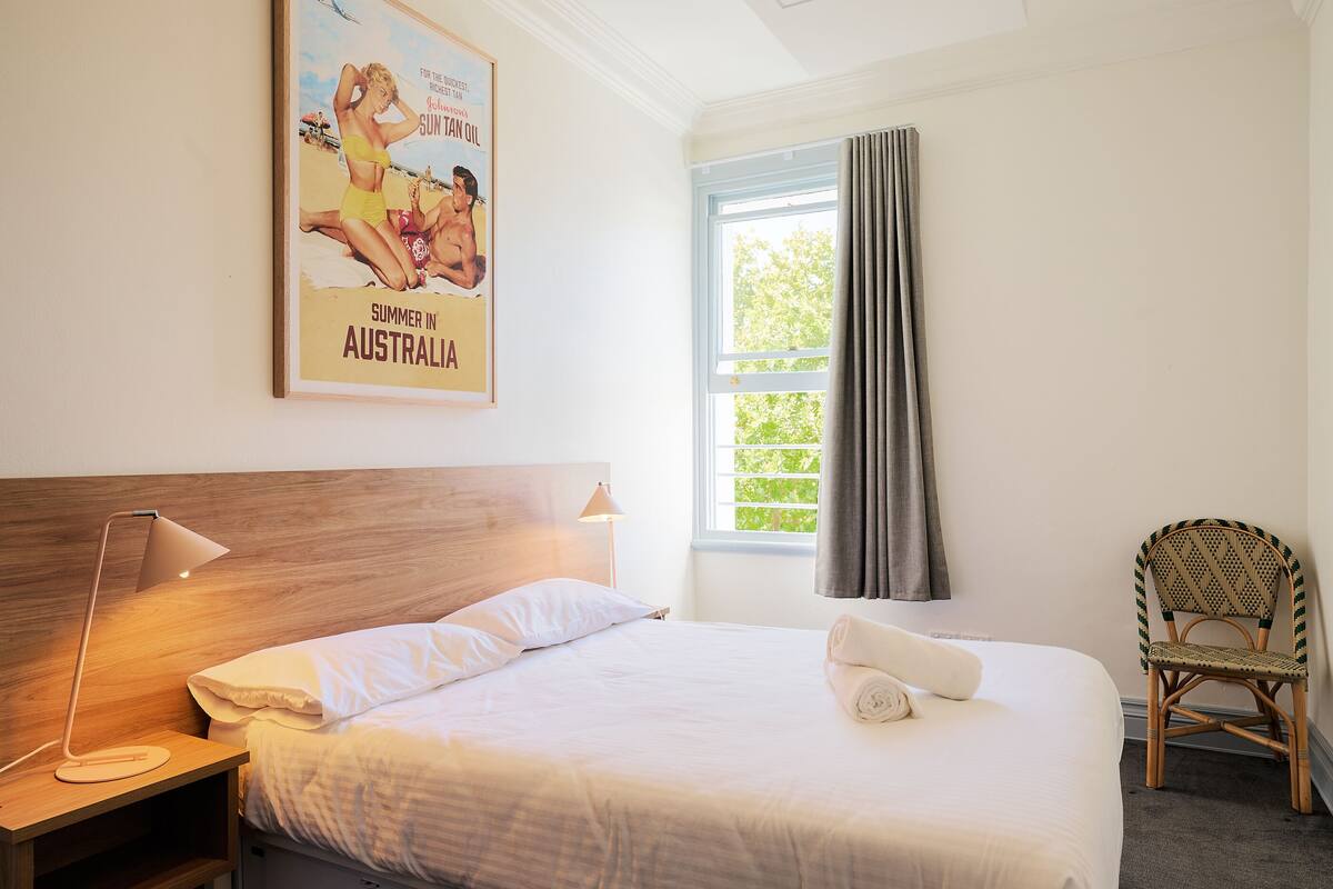 A neatly made bed with white linens, a wooden headboard, a nightstand with a lamp, a wicker chair, and a window with curtains.