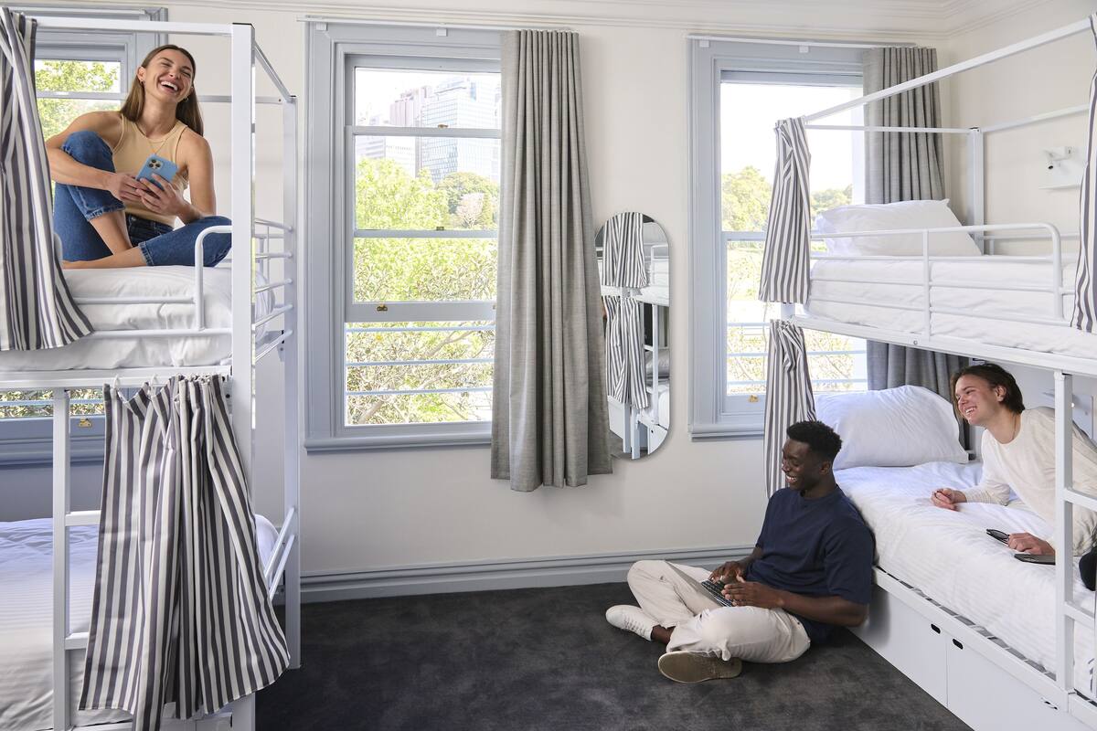 A dormitory room with bunk beds, a large window with curtains, and a mirror on the wall.