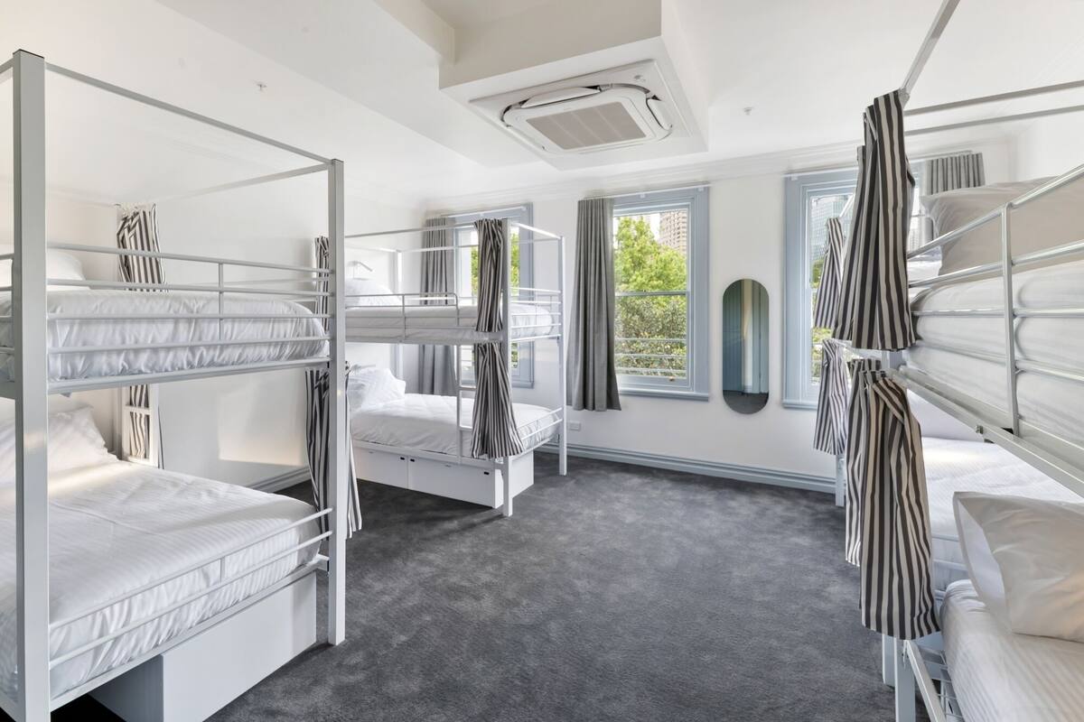 A dormitory room with bunk beds, a large window, and a ceiling-mounted air conditioning unit.