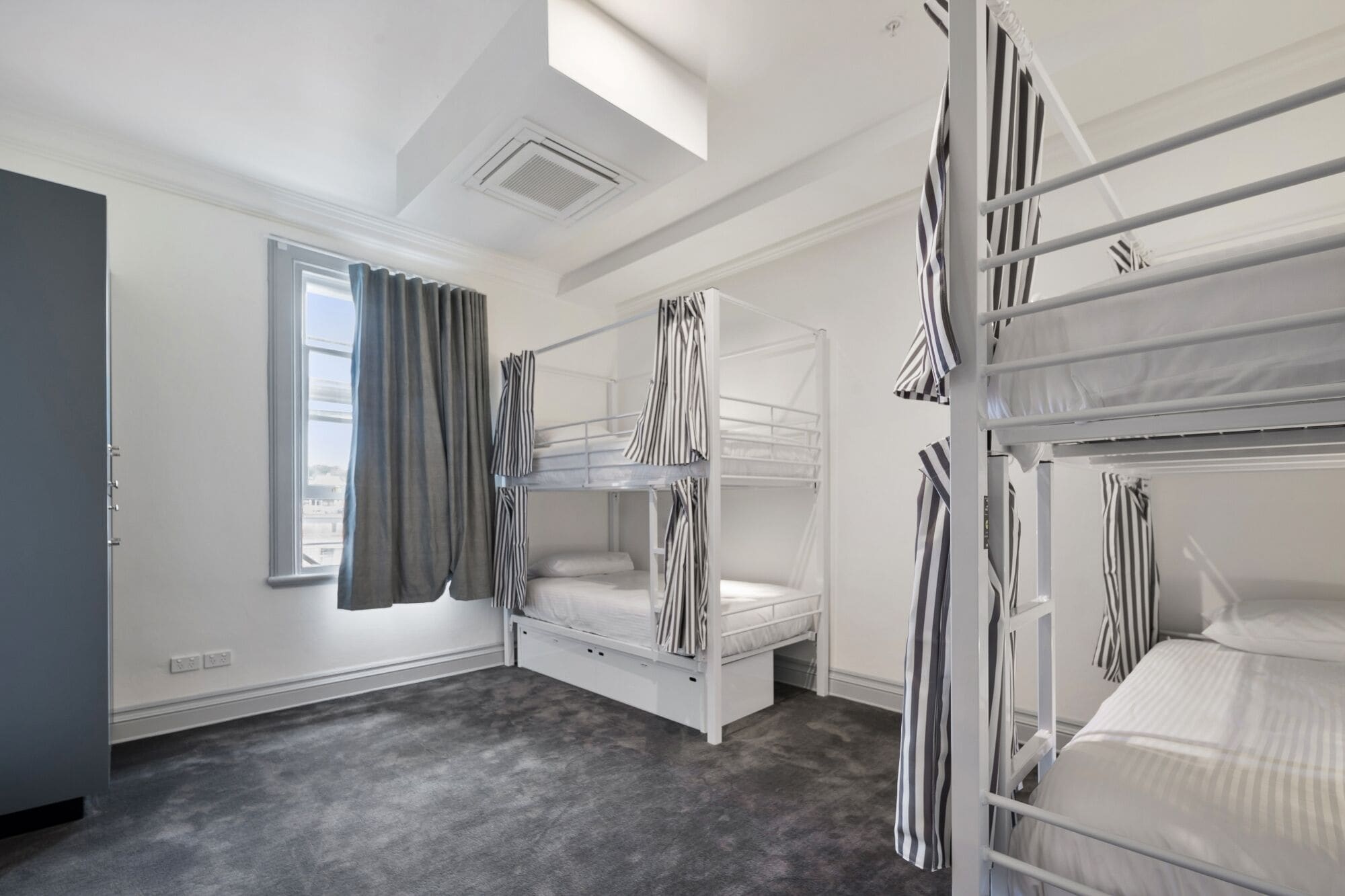 A room with bunk beds, a window with curtains, a ceiling vent, and a grey carpet.