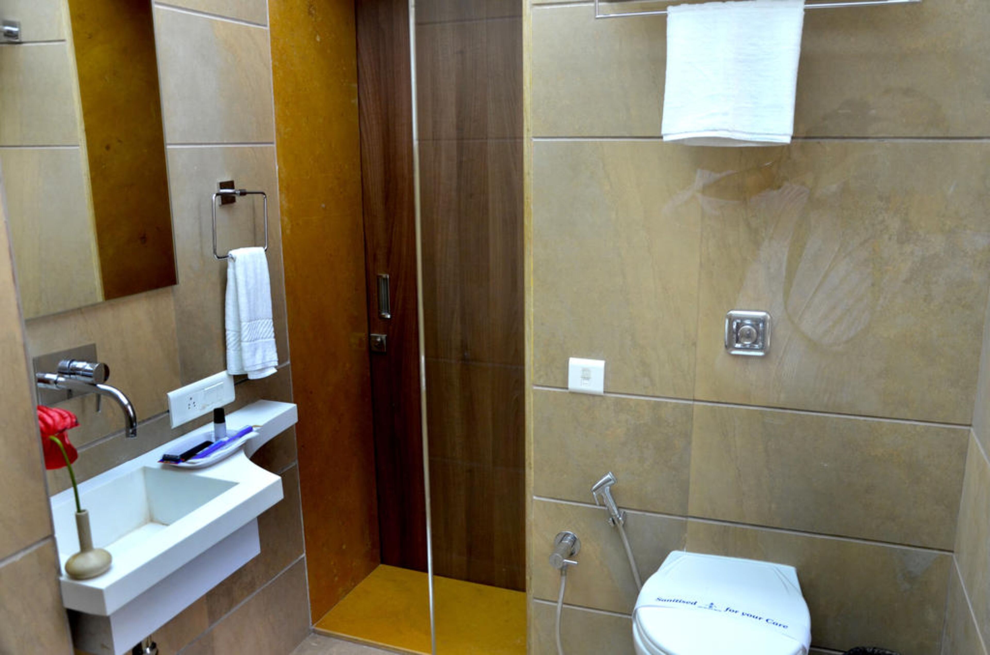 A bathroom with a white sink, mirror, and toilet. There is a shower area with a glass door and a towel hanging on the wall.