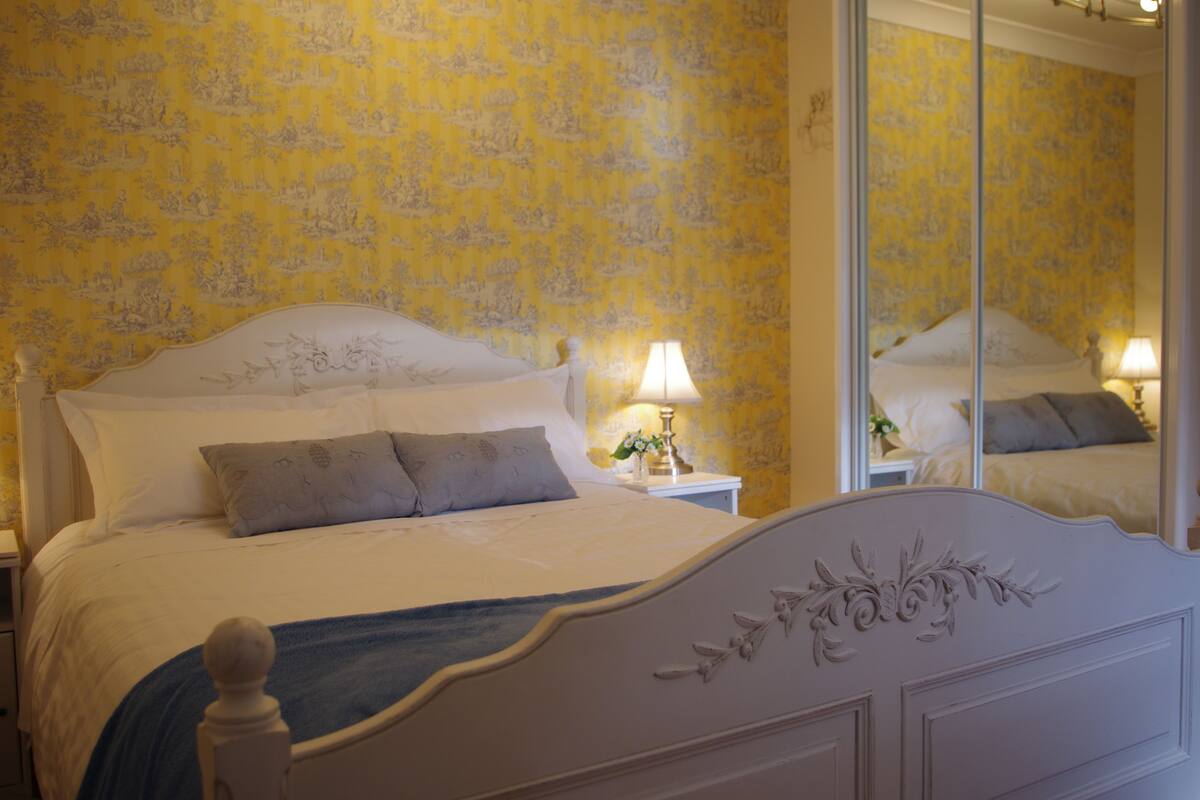 A neatly made bed with a white headboard, a blue throw blanket, and a nightstand with a lamp.