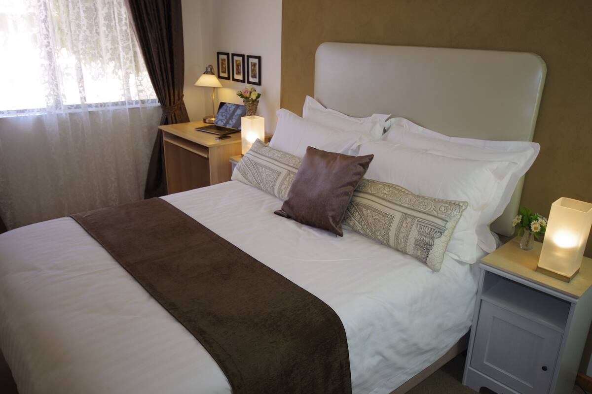 A neatly made bed with a brown throw blanket, white bedding, and decorative pillows. There is a bedside table with a lamp, a laptop, and a vase with flowers. The room has a window with sheer curtains and a framed picture on the wall.
