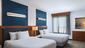 Une chambre d’hôtel avec deux lits, une table de chevet, une lampe, une tête de lit en bois, une fenêtre avec des rideaux et un t éléviseur fixé au mur.