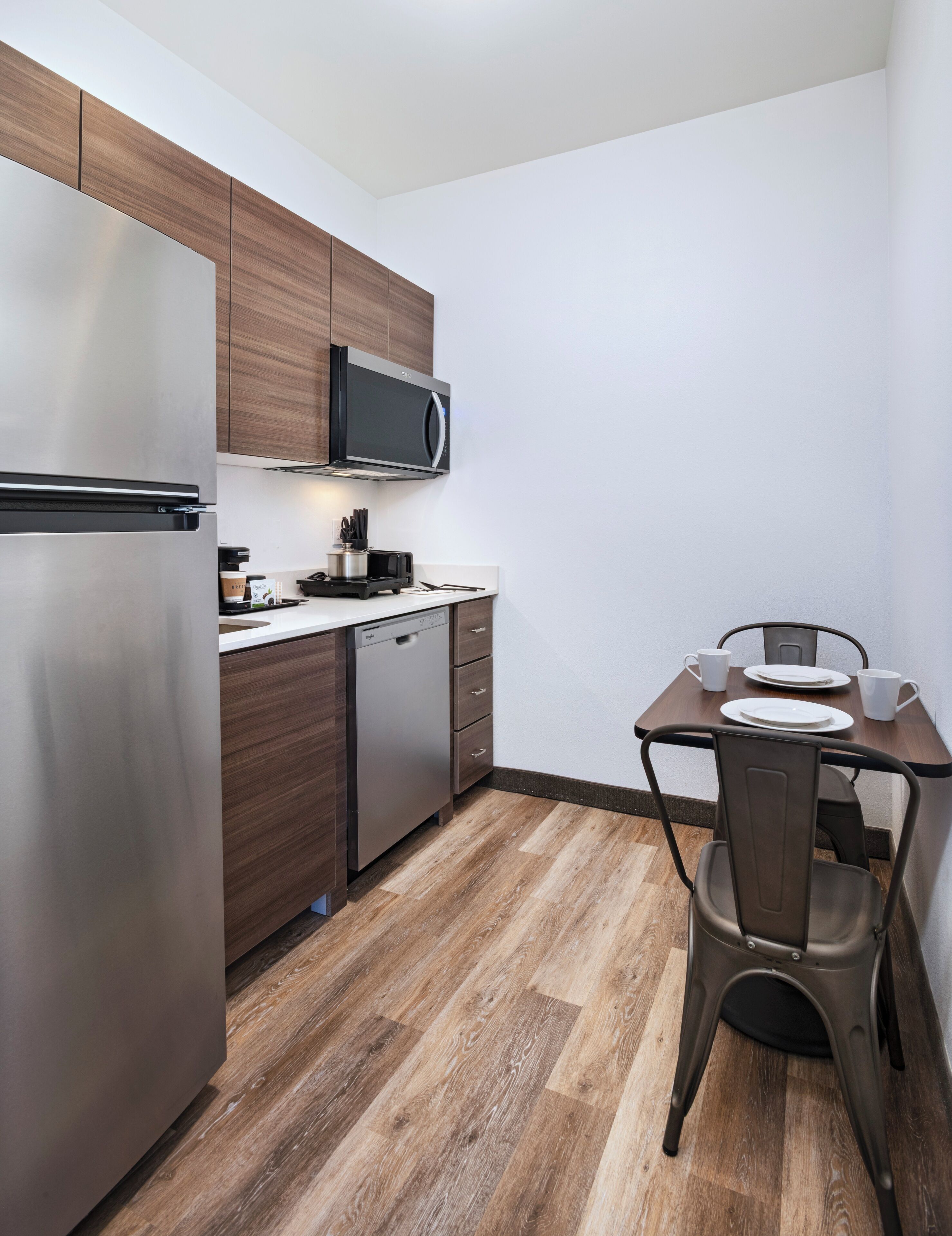 A compact kitchen with a stainless steel refrigerator, a microwave, a dishwasher, and a dining table with chairs.